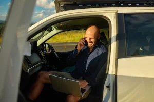 A tradesman sits in his car, working on a laptop and talking on the phone, with a relaxed expression.